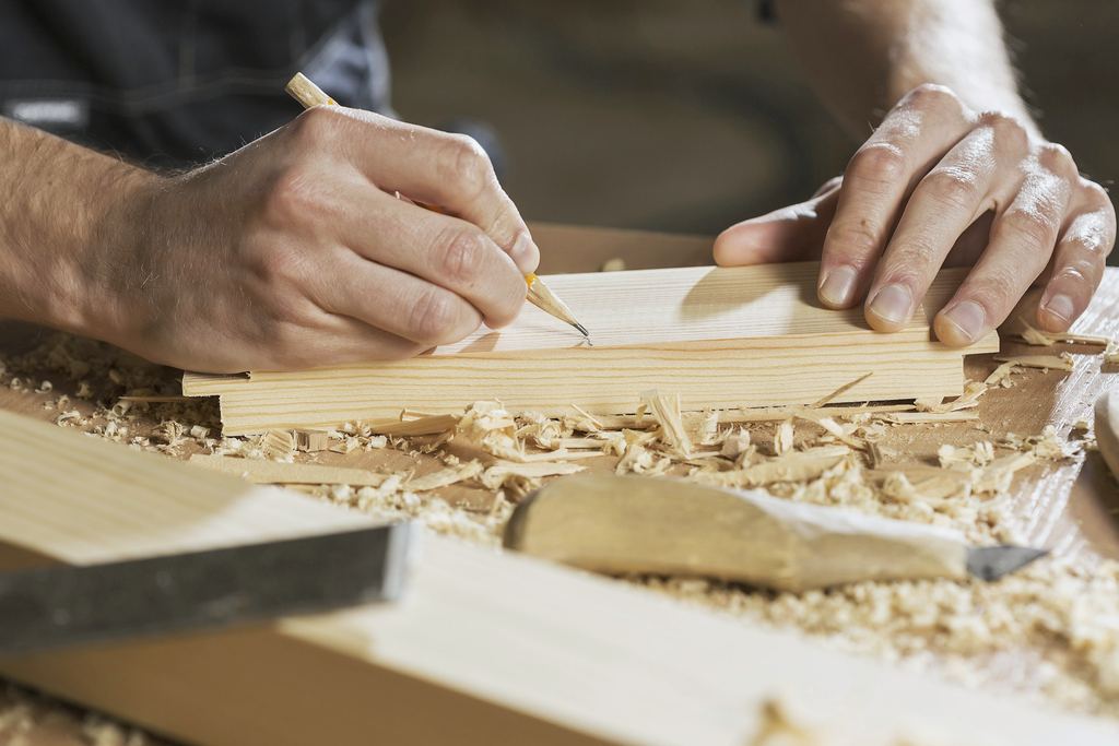 Eine Hand zeichnet mit einem Bleistift eine Linie auf ein helles Holzstück, daneben Hobelspäne und ein Stemmeisen.