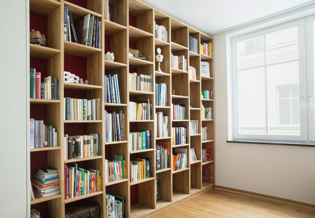 Raumhohe Bücherwand aus hellem Holz mit vielen gleich großen Fächern, rechts ein weißes Sprossenfenster.