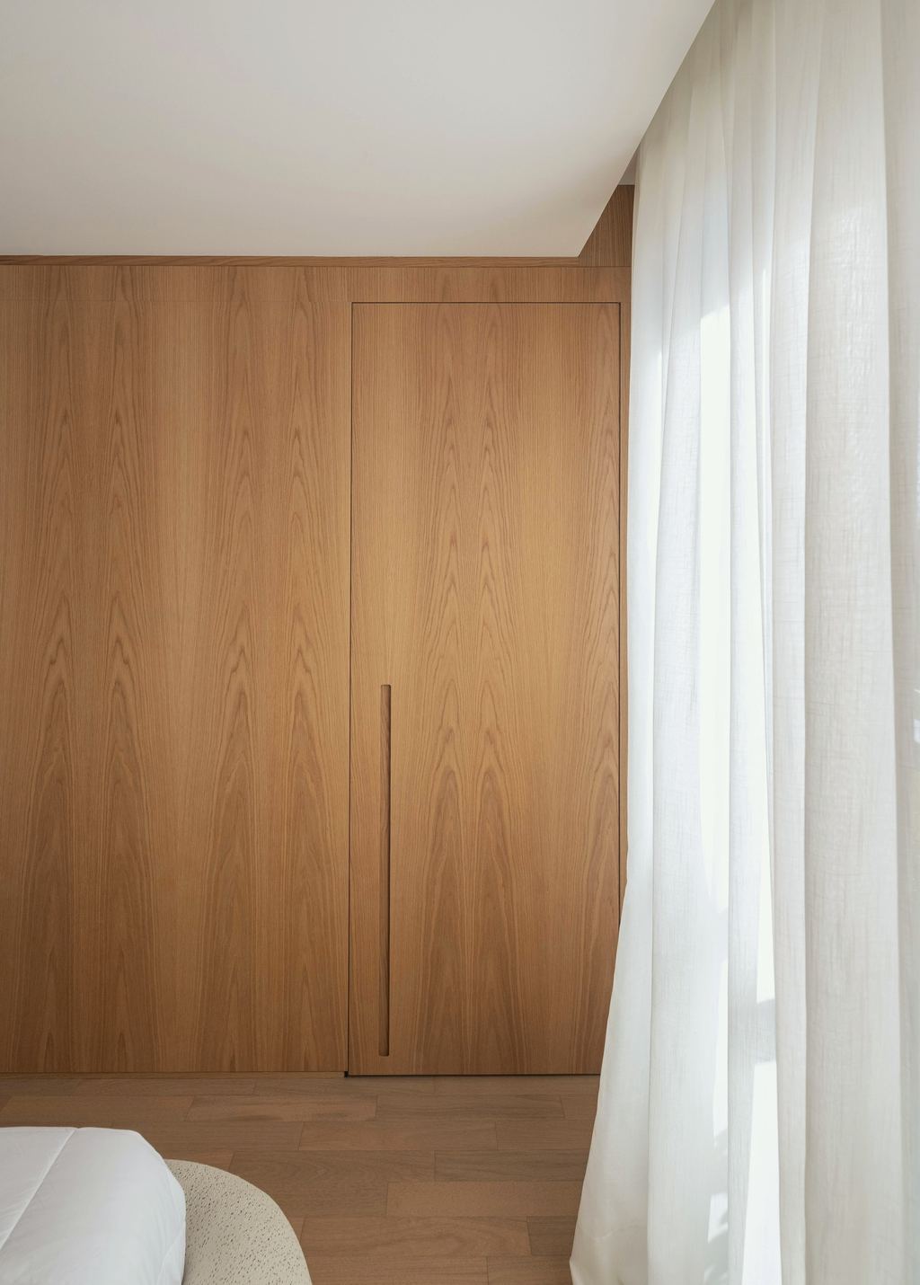 Raumhoher Einbauschrank aus Eiche mit glatten Fronten und schmaler senkrechter Griffmulde, daneben ein Fenster mit hellem Vorhang.