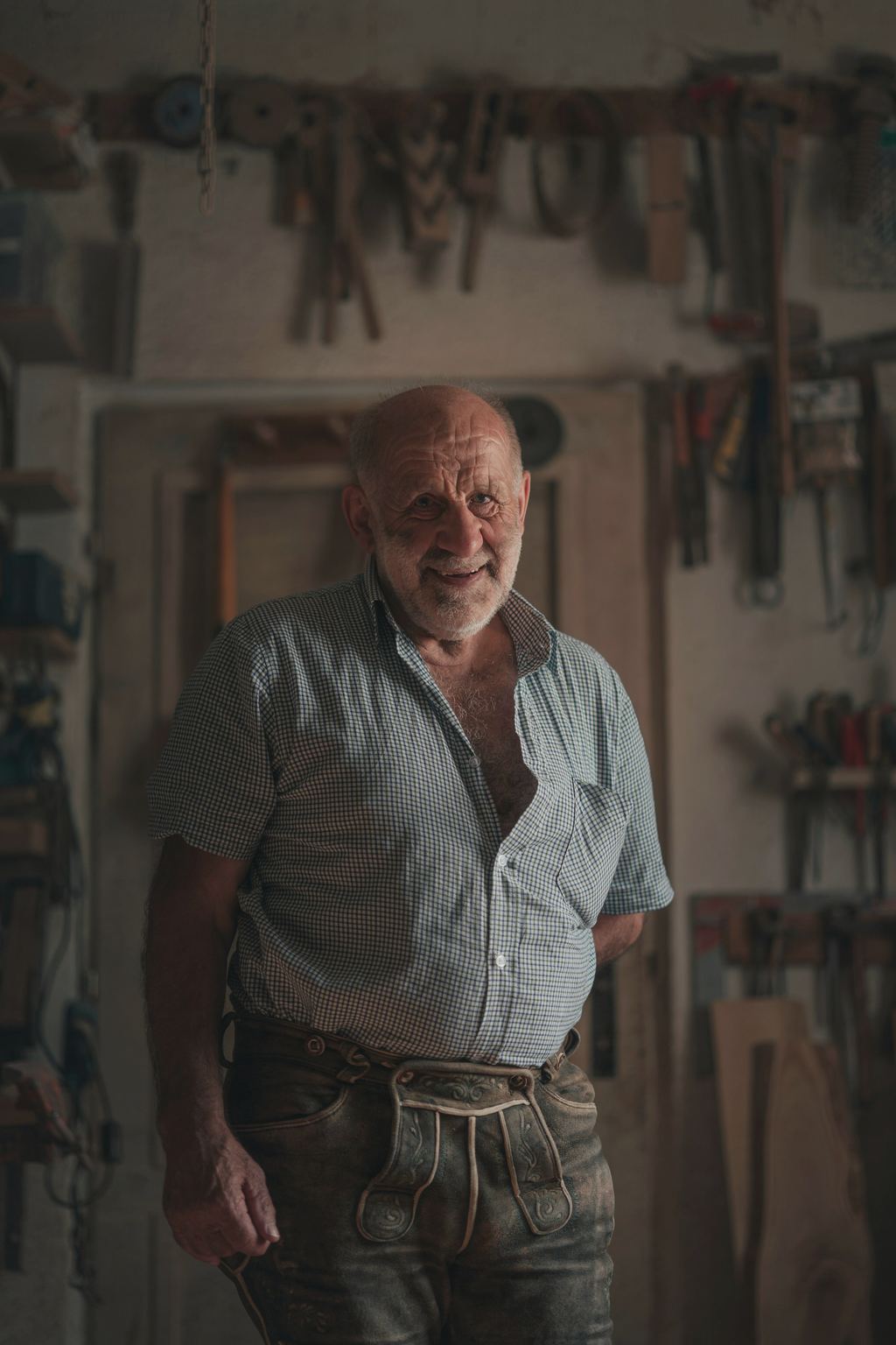 Ein älterer Mann in Lederhose und Hemd steht lächelnd vor einer Wand mit Werkzeug, Symbolfoto.