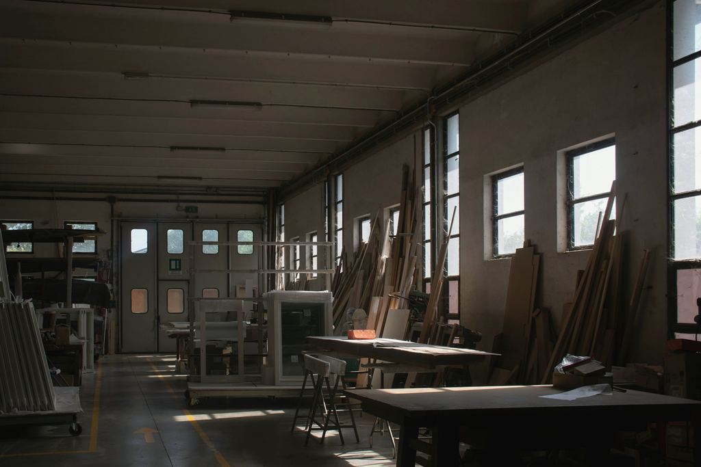 Große Werkstatthalle mit Fensterreihe, Werkbänken und an die Wand gelehnten Brettern.