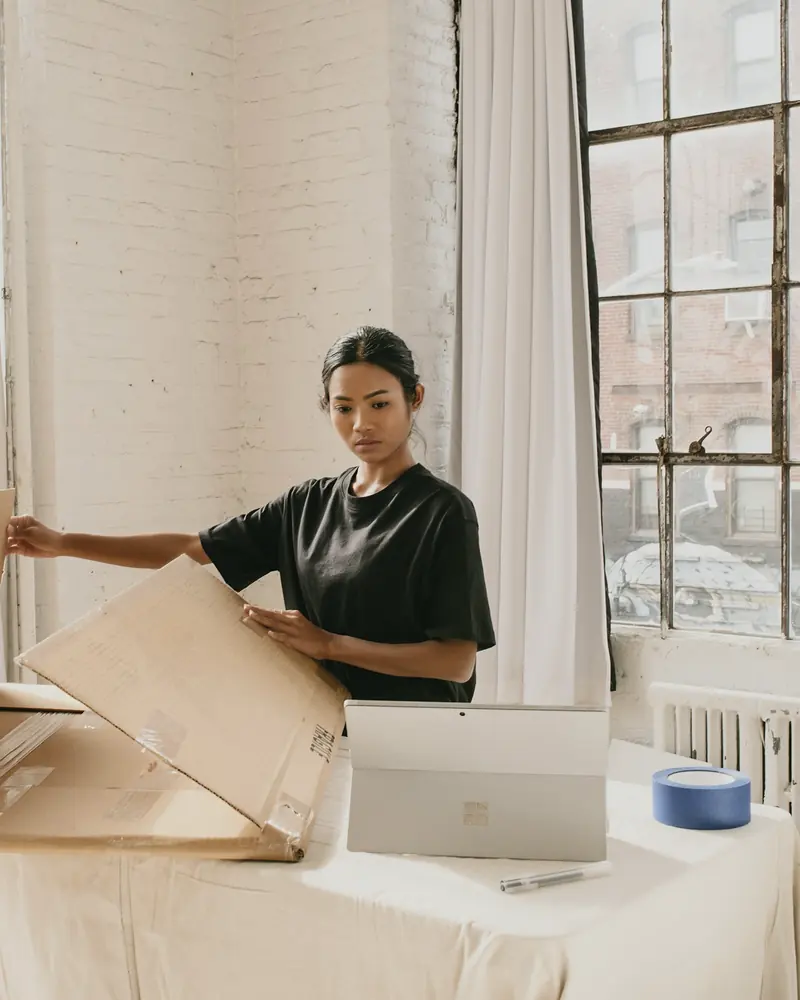 Eine Frau faltet in einem hellen Atelier einen großen Karton auf, daneben ein Laptop.